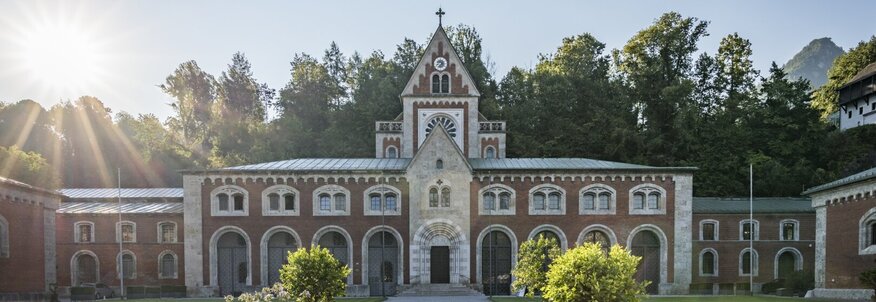 Das große rote Gebäude aus Backstein ist die Alte Saline in Bad Reichenhall. Hier wird seit langem salzhaltige AlpenSole gefördert. Die Saline ist ein Industriedenkmal und das Salzmuseum ist in ihr untergebracht. Im Vordergrund ist ein schöner Park mit geschotterten Wegen und grünem Rasen zu sehen. Hinter dem Gebäude stehen Bäume und die Sonne geht gerade am blauen Himmel über der Alten Saline auf.