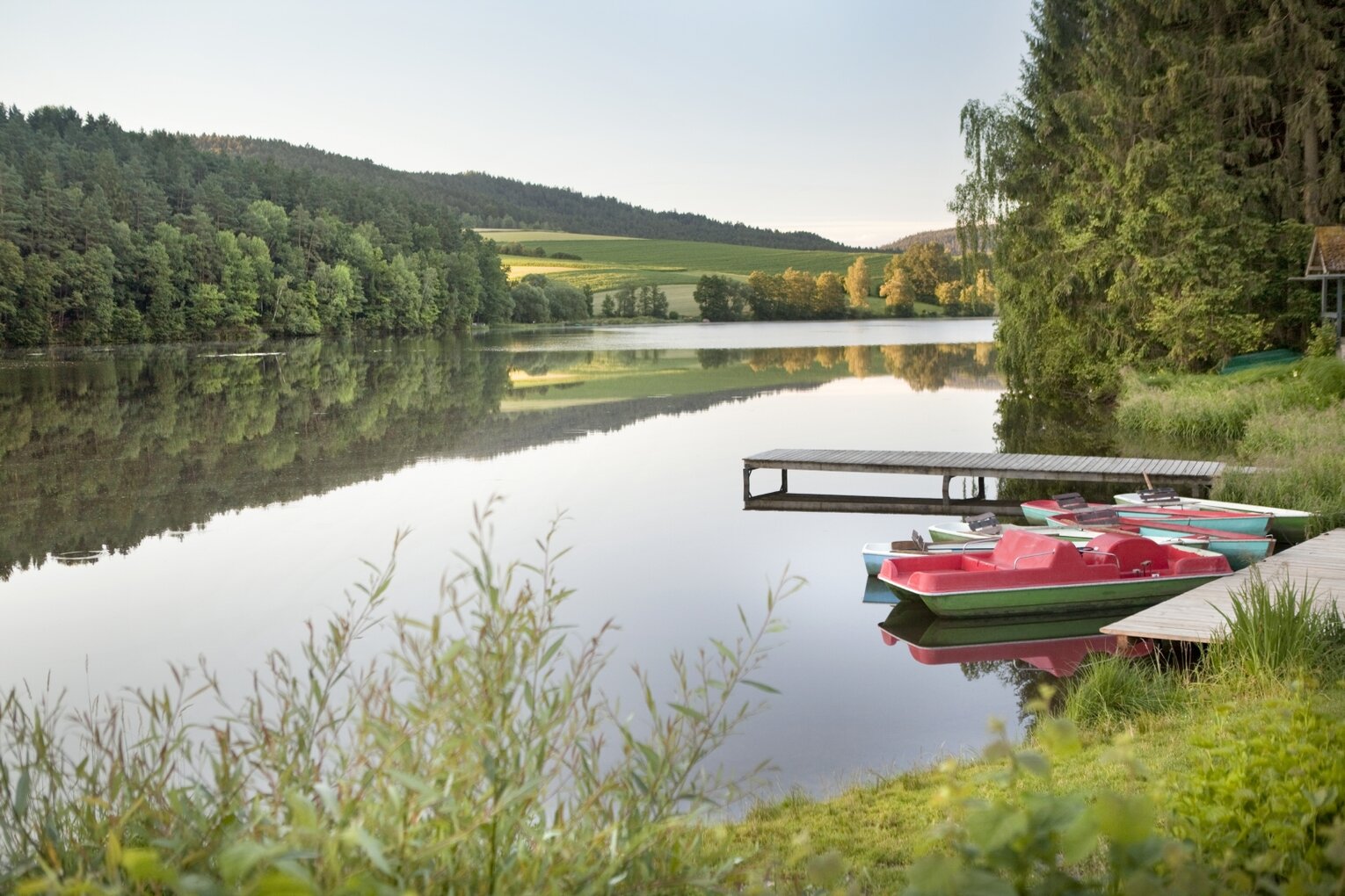 Hier sehen Sie eine See, welcher von Bäumen, Büschen und Gräsern umgeben ist. Ein kleiner Steg führt in den See, am Ufer sind kleine Tretboote zu sehen.