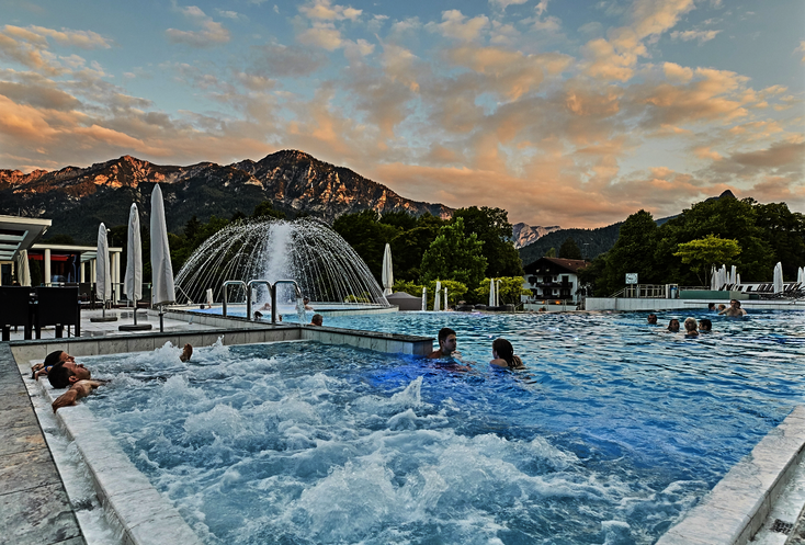 Hier sehen Sie eine Pool im Außenbereich. Es sind vereinzelnt Menschen in dem Pool und liegen auf den Massagedüsen. Im Hintergrund sind Berge zu sehen. 