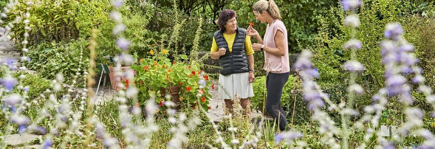 Ich sehe zwei Frauen, die sich in einem Kräutergarten unterhalten. Die Linke Frau trägt einen weißen Rock, ein gelbes T-shirt  und drüber eine schwarze Weste. Die rechte Frau trägt eine schwarze Sporthose und ein rosa Oberteil. 