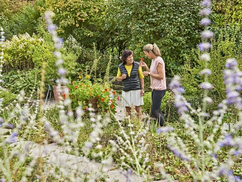 Ich sehe zwei Frauen, die sich in einem Kräutergarten unterhalten. Die Linke Frau trägt einen weißen Rock, ein gelbes T-shirt  und drüber eine schwarze Weste. Die rechte Frau trägt eine schwarze Sporthose und ein rosa Oberteil. 