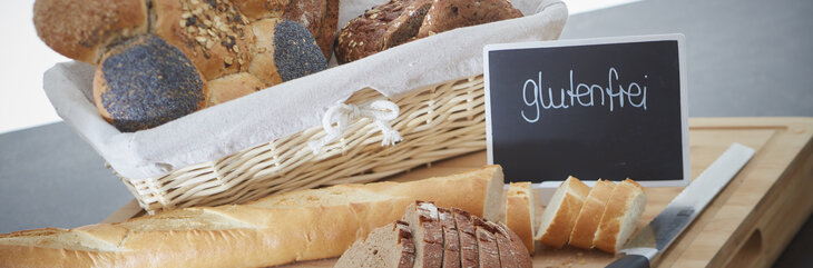 Hier sehen Sie einen angerichteten Brotkorb, sowie ein Schneidebrett und ein Brotmesser. Daneben steht auf einem kleinen Schild "glutenfrei". 