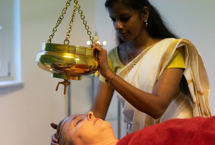 Hier sehen Sie eine Frau, welche auf einer Lieg ligt. Ein Therapeutin steht neben ihr und macht eine ayurvedische Öl Massage. 