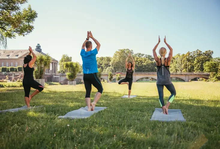 Hier sehen Sie mehrere Frauen auf einer Wiese stehen. Sie haben Yogamatten unter ihren Füßen und praktizieren Yoga. 