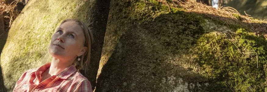 Hier sehen Sie eine Dame, welche sich an einem moosigen Stein im Wald anlehnt. Sie blickt in den Himmel rauf.