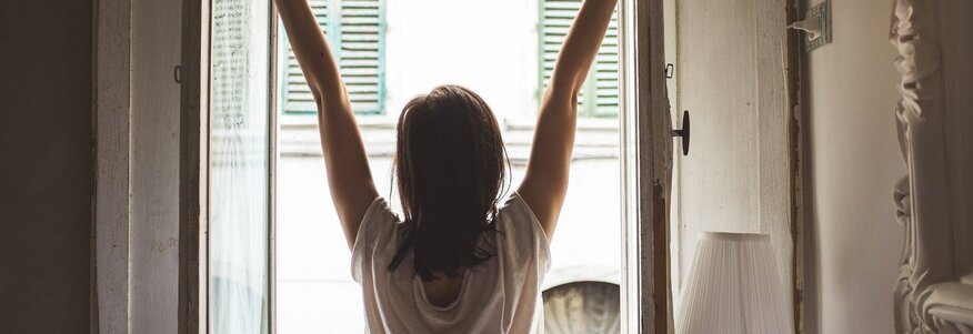 Hier sehen Sie eine Frau die sich vor einem offenen Fenster streckt. Sie trägt noch einen Schlafanzug und ist mit dem Rücken zu uns gedreht. 