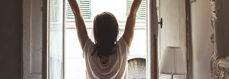 Hier sehen Sie eine Frau die sich vor einem offenen Fenster streckt. Sie trägt noch einen Schlafanzug und ist mit dem Rücken zu uns gedreht. 