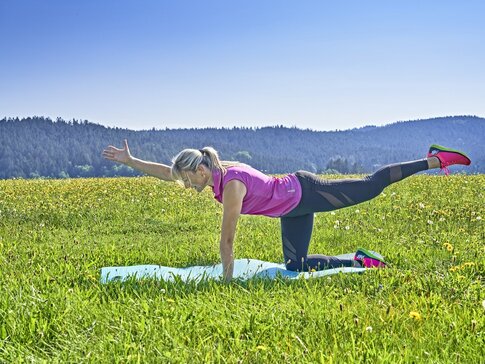 Hier sehen Sie die GESUNDES BAYERN Expertin Jana Schäfer-Schalk auf einer Wiese. Sie macht auf einr Sportmatte Gymnastikübungen. 