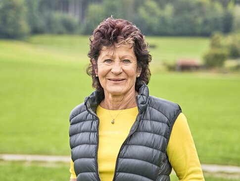 Sie sehen ein Bild von Ines Wurm-Fenkl, der GESUNDES BAYERN-Expertin aus Bad Wörishofen. Frau Wurm-Fenkl trägt ein gelbes Oberteil mit einer schwarzen Steppweste darüber. Sie hat dunkle lockige Haare und lächelt in die Kamera. Im Hintergrund sind Wiesen und Wald zu sehen.