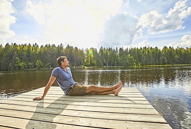 Hier sehen Sie einen Steg der raus auf einen See führt. Ein Mann sitzt mit ausgestreckten beinen seitlich zu uns gedreht am Ende des Stegs. Hinter dem See sind Bäume zu sehen. 