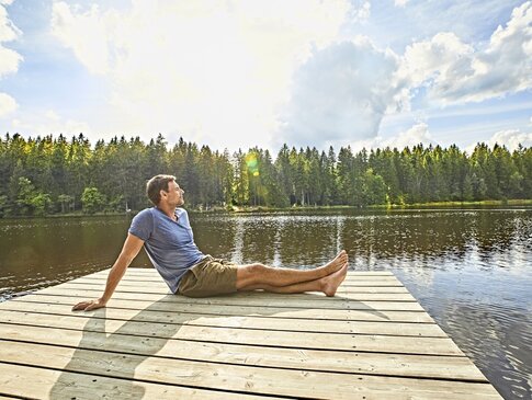 Hier sehen Sie einen Steg der raus auf einen See führt. Ein Mann sitzt mit ausgestreckten beinen seitlich zu uns gedreht am Ende des Stegs. Hinter dem See sind Bäume zu sehen. 