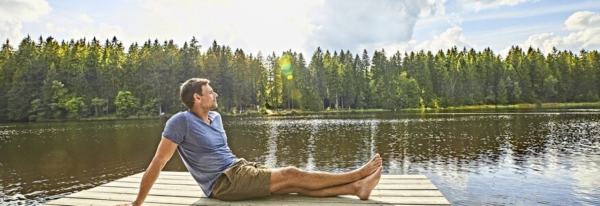 Hier sehen Sie einen Steg der raus auf einen See führt. Ein Mann sitzt mit ausgestreckten beinen seitlich zu uns gedreht am Ende des Stegs. Hinter dem See sind Bäume zu sehen. 