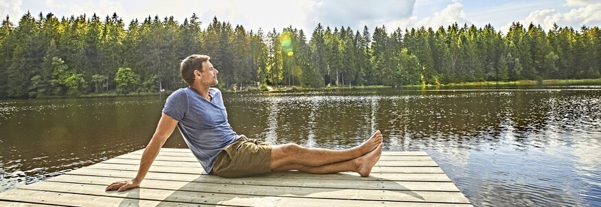 Hier sehen Sie einen Steg der raus auf einen See führt. Ein Mann sitzt mit ausgestreckten beinen seitlich zu uns gedreht am Ende des Stegs. Hinter dem See sind Bäume zu sehen. 