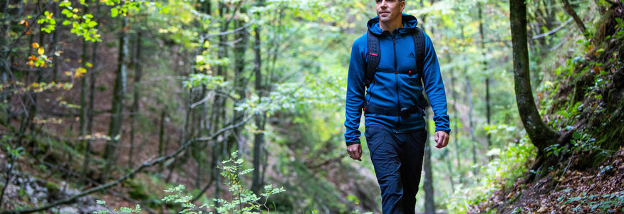 Hier sehen Sie den ehemaligen Profifußballer Philipp Lahm in Wanderbekleidung. Er spaziert durch eine Waldweg. Um ihm herum sind Büume un Büsche zu sehen, auf dem Boden liegt trockenes Laub. 