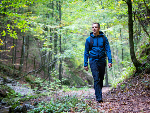 Hier sehen Sie den ehemaligen Profifußballer Philipp Lahm in Wanderbekleidung. Er spaziert durch eine Waldweg. Um ihm herum sind Büume un Büsche zu sehen, auf dem Boden liegt trockenes Laub. 