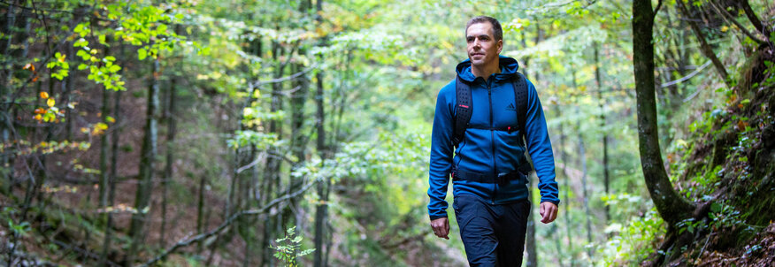 Hier sehen Sie den ehemaligen Profifußballer Philipp Lahm in Wanderbekleidung. Er spaziert durch eine Waldweg. Um ihm herum sind Büume un Büsche zu sehen, auf dem Boden liegt trockenes Laub. 