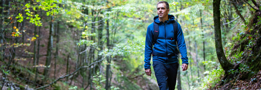 Hier sehen Sie den ehemaligen Profifußballer Philipp Lahm in Wanderbekleidung. Er spaziert durch eine Waldweg. Um ihm herum sind Büume un Büsche zu sehen, auf dem Boden liegt trockenes Laub. 