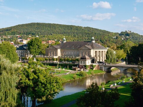 Zu sehen ist ein großes Gebäude, der Regentenbau in Bad Kissingen. Im Hintergrund ist ein Berg mit vielen Bäumen zu sehen, vor dem Gebäude fließt ein kleiner Fluss, die Fränkische Saale vorbei. Am Flussufer stehen auch Bäume.