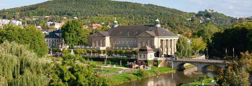 Zu sehen ist ein großes Gebäude, der Regentenbau in Bad Kissingen. Im Hintergrund ist ein Berg mit vielen Bäumen zu sehen, vor dem Gebäude fließt ein kleiner Fluss, die Fränkische Saale vorbei. Am Flussufer stehen auch Bäume.