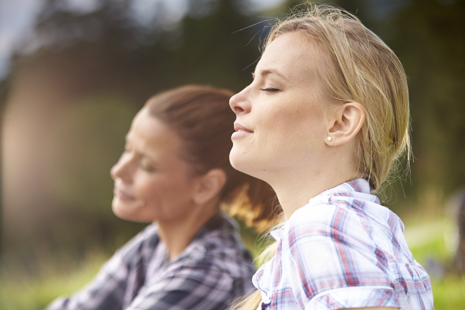 Hier sehen Sie im Vordergrund zwei Frauen in der Natur, die mit geschlossenen Augen tief durchatmen. Die Sonne scheint auf die Frauen; im Hintergrund sind unscharf Bäume und Wiese zu erkennen.