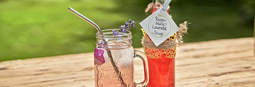 Hier sehen Sie ein Glas und eine Flasche mit einem Etikett und einem rosa Inhalt auf einem Holztisch in der Natur stehen. 