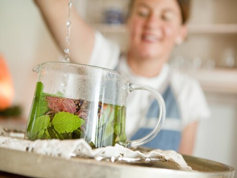 Hier sehen Sie eine Teekanne aus Glas in der viele frische Kräuter sind. Eine Frau im Hintergrund ist unscharf zu sehen. Sie schüttet heißes Wasser in die Teekanne und brüht den Tee auf. 