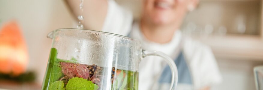 Hier sehen Sie eine Teekanne aus Glas in der viele frische Kräuter sind. Eine Frau im Hintergrund ist unscharf zu sehen. Sie schüttet heißes Wasser in die Teekanne und brüht den Tee auf. 