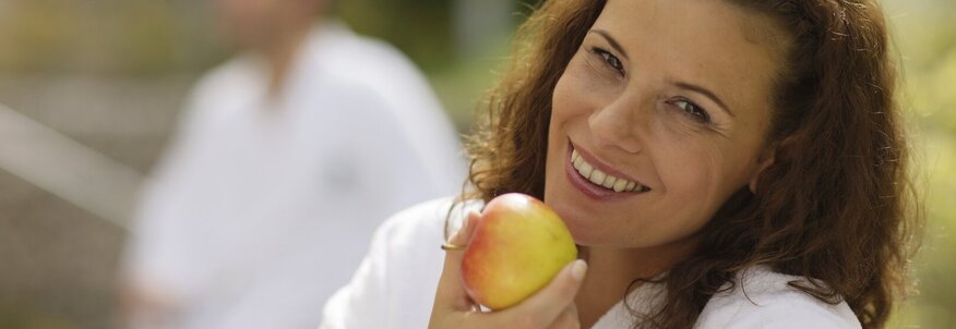 Hier sehen Sie eine Frau in einem Bademantel, sie lacht in die Kamera und hat einen Apfel in der Hand. 