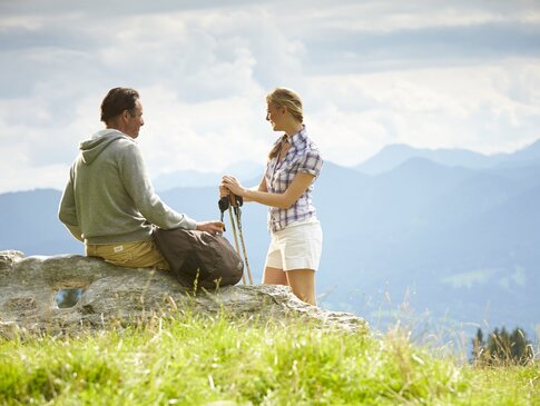 Hier sehen Sie ein Paar, welches im Sommer in den Bergen wandern ist. Der Mann sitzt auf einem Felsen und hat den Rucksack abgelegt. Die Frau steht daneben und stützt sich an den Nordicwalking Stöcken ab. Vor ihnen ist ein Bergpanorama zu sehen. 