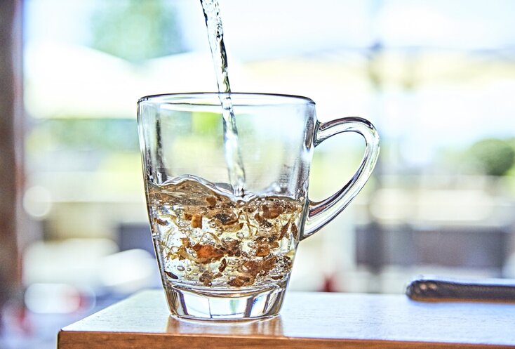 Hier sehen Sie eine Tasse aus Glas mit getrockneten Kräutern. Es wird Wasser aufgegossen. 