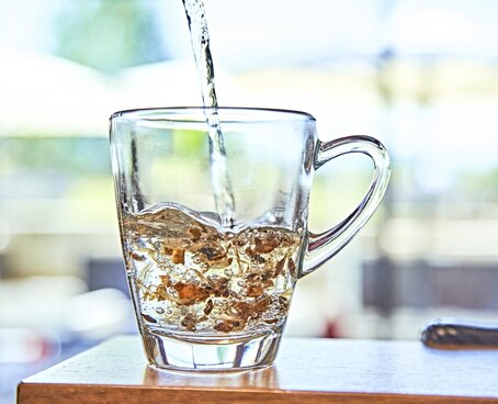 Hier sehen Sie eine Tasse aus Glas mit getrockneten Kräutern. Es wird Wasser aufgegossen. 