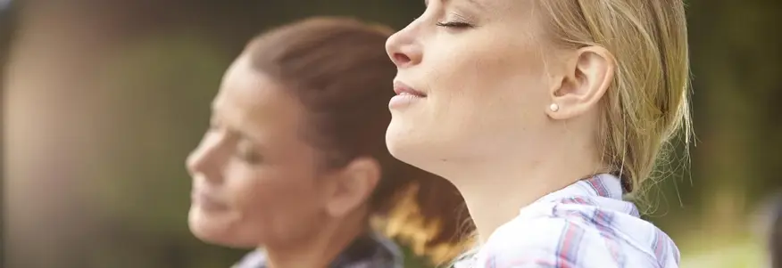 Nahaufnahme von zwei Frauen, die in einer Wiese sitzen und tief durchatmen.