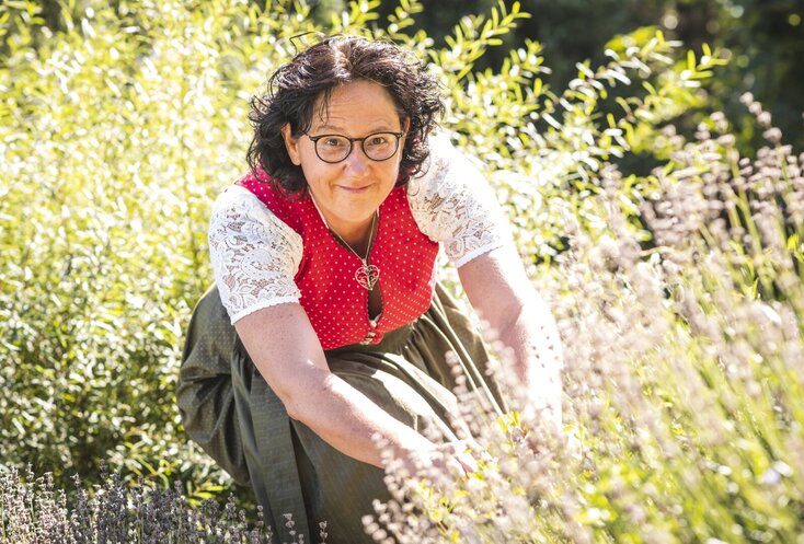 Hier sehen Sie eine Frau mit dunklen, gelockten Haaren, welche ein Dirndl trägt und Kräuter sammelt.