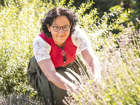 Hier sehen Sie eine Frau mit dunklen, gelockten Haaren, welche ein Dirndl trägt und Kräuter sammelt.