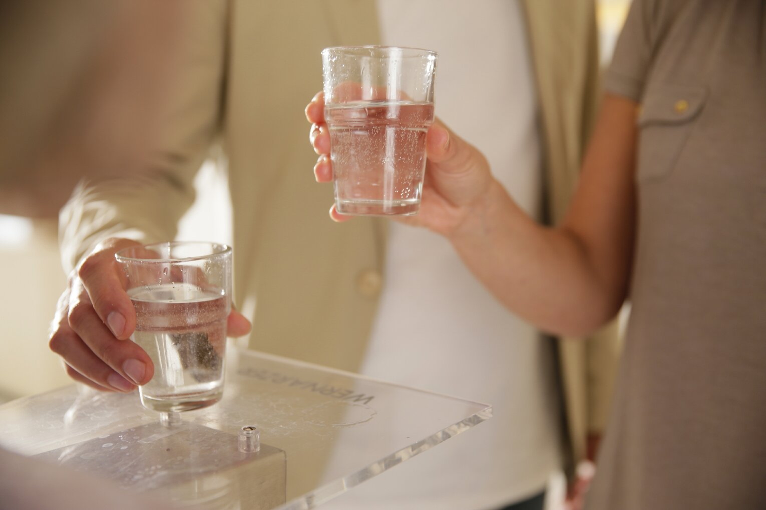 Hier sehen Sie den Oberkörper einer Frau und eines Mannes. Beide halten in ihrer Hand ein Glas mit Heilwasser gefüllt. 