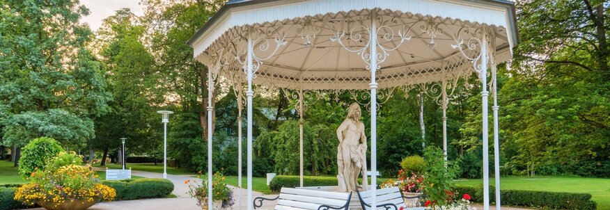 Sie sehen einen historischen Pavillon im Kurpark von Bad Steben. Im Pavillon stehen Bänke, damit man sich ausruhen kann und eine große Steinfigur.