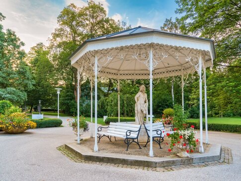 Sie sehen einen historischen Pavillon im Kurpark von Bad Steben. Im Pavillon stehen Bänke, damit man sich ausruhen kann und eine große Steinfigur.