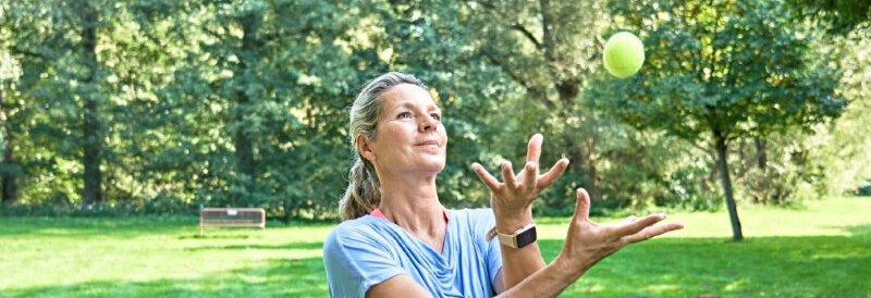 Hier sehen Sie eine Frau mit blondem Haar und blauem T-Shirt, welche eine Übung mit zwei Bällen ausübt.