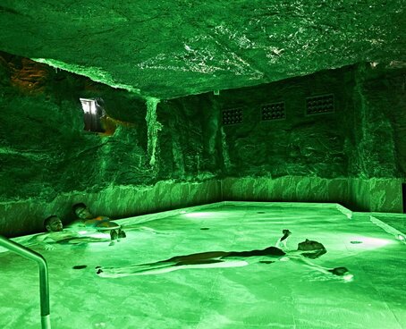 Hier sehen Sie ein Solebad im Innenbereich eines Kurhauses. Eine Frau schwimmt mit dem Rücken auf der Oberfläche das salzhaltigen Wassers. Der Raum ist dunkel, niedrig und grün beleuchtet. 