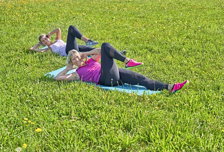 Hier sehen Sie zwei Frauen auf einer Wiese. Sie tragen Sportkleidung und machen Bauchübungen auf dem Boden.