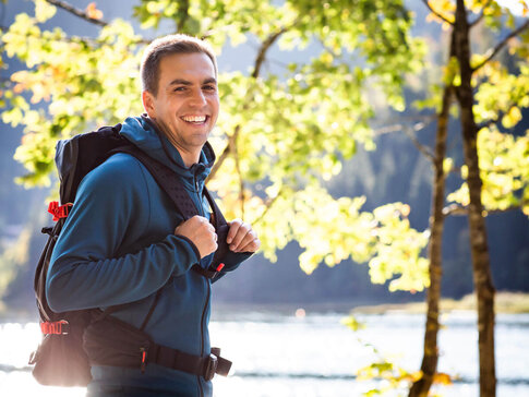 Hier sehen Sie den ehemaligen Profi-Fußballer Philipp Lahm. In Wanderbekleidung steht er an einem See zwischen Bäumen. Er schaut in richtung uns und lächelt. 