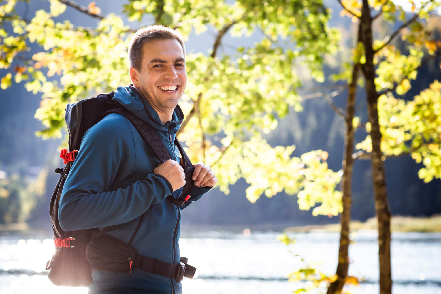 Hier sehen Sie den ehemaligen Profi-Fußballer Philipp Lahm. In Wanderbekleidung steht er an einem See zwischen Bäumen. Er schaut in richtung uns und lächelt. 