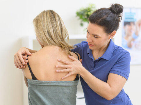 Hier sehen Sie eine Frau die mit dem Rücken zu uns sitzt und ein Handtuch umgewickelt hat. Eine Therapeutin steht neben ihr und behandelt den Rücken der Patientin. 