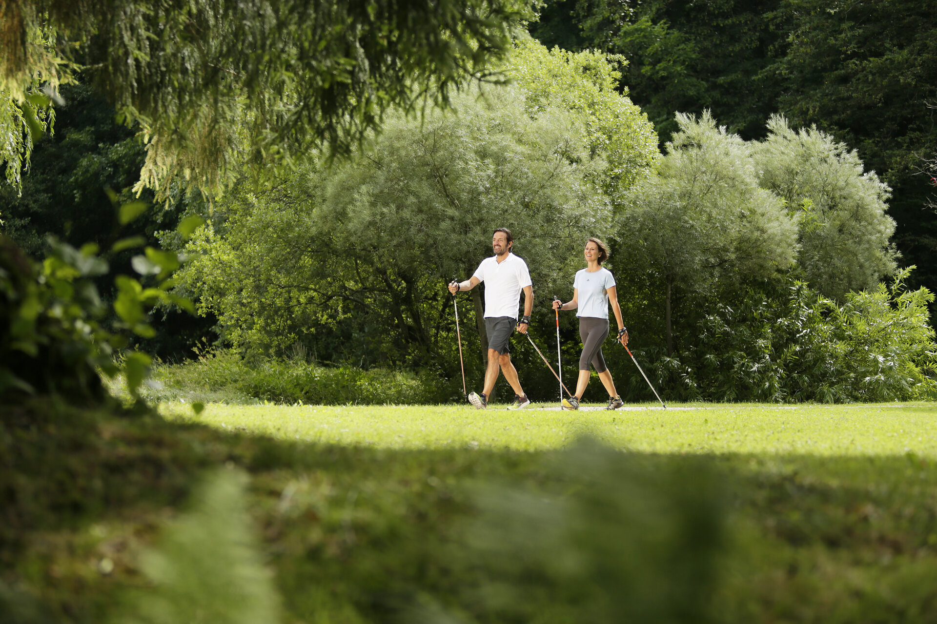 Hier sehen Sie zwei Personen durch den Kurgarten Bad Brückenau spazieren. Beide Personen haben Nordic Walking Stöcke dabei. 