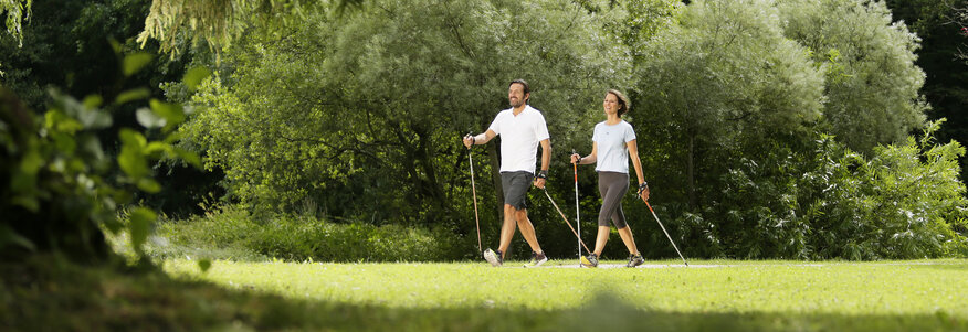 Hier sehen Sie zwei Personen durch den Kurgarten Bad Brückenau spazieren. Beide Personen haben Nordic Walking Stöcke dabei. 